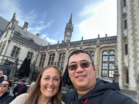 Smiling couple taking a selfie in front of a historic neo-gothic building with a clock tower under a partly cloudy sky.