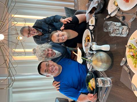 Four diners gathered around a restaurant table enjoying plated meals and smiling at the camera.