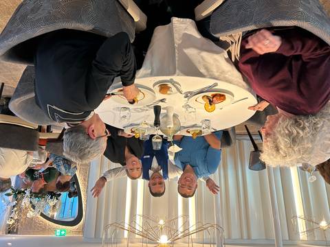 Group of travelers and a waiter cheerfully posing around a round table set with lunch dishes.
