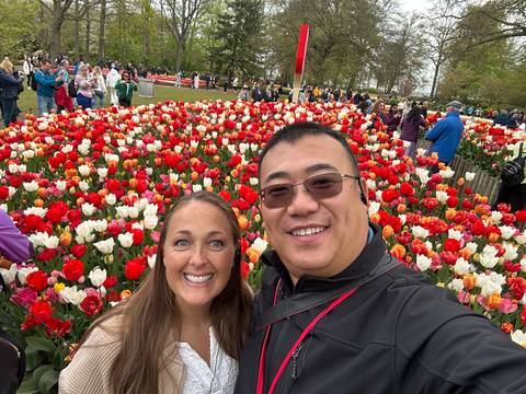 Smiling couple surrounded by vibrant rows of tulips in a crowded flower garden.