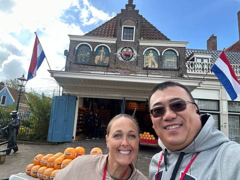 Couple smiling in front of a Dutch cheese shop with national flags and stacked cheese wheels.