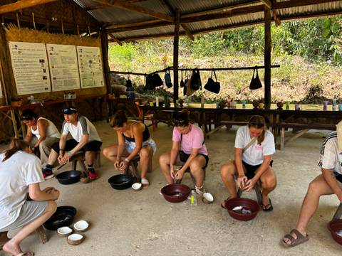 Small group of travellers seated under a thatched shelter learning to grind coconut using bowls.