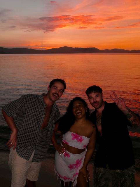 Three friends posing and laughing in front of vivid orange sunset over calm sea