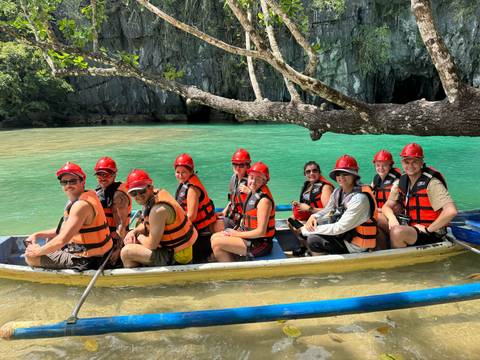 Tour group wearing life vests and red helmets seated in paddle boat on turquoise river emerging from limestone cave