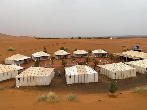 Neat rows of white canvas tents form a desert camp set amid reddish sand with distant dunes.