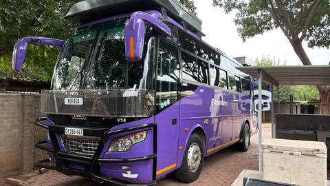 Daytime view of a purple expedition bus parked on a paved lot beside trees.