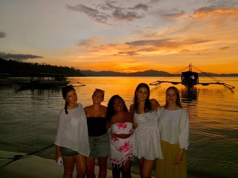 Five young women pose together on a seaside promenade, a traditional outrigger boat and vibrant orange sunset reflecting on calm tropical water behind them.