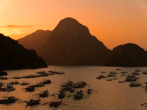 Orange sunset over a sheltered bay filled with outrigger boats and a dramatic silhouetted limestone mountain.