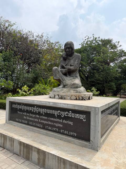 A sombre genocide memorial statue of a mother and child sits atop a stone plinth inscribed with remembrance text.