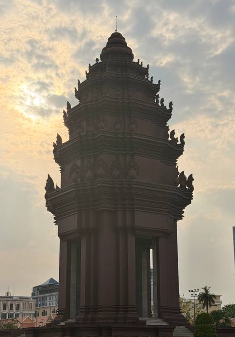 A silhouetted monument tower with ornate naga corners rises into a hazy sky with the sun glowing behind it.