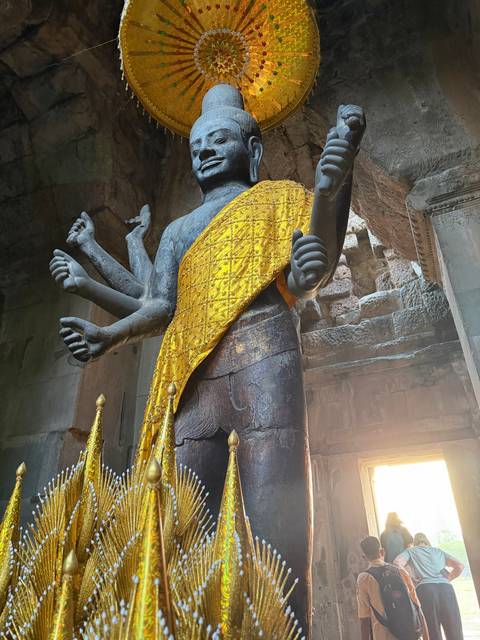 A multi-armed stone Vishnu statue draped in a golden cloth stands inside the dim corridors of Angkor Wat.