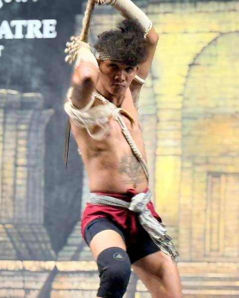 A traditional Cambodian male dancer mid-performance wearing red costume under stage lighting appears slightly motion-blurred.