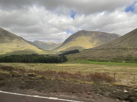 Sunlit Scottish highland valley with rolling hills, distant forests and dramatic cloud shadows.
