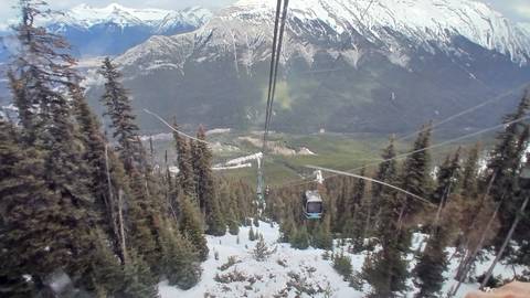 Gondola car descends over snowy alpine forest toward a valley and rugged peaks.