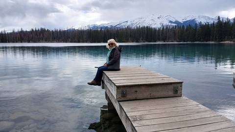 Woman sits quietly on a wooden dock extending into a glassy alpine lake surrounded by forested peaks.