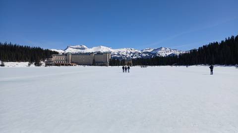 Frozen lake with grand hotel and snow-covered Rocky Mountains under a clear blue sky; tiny figures walk across the ice.