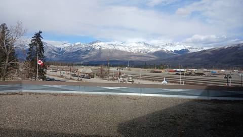 Rail yard and Canadian flag with a backdrop of snow-clad Rockies under partly cloudy sky.