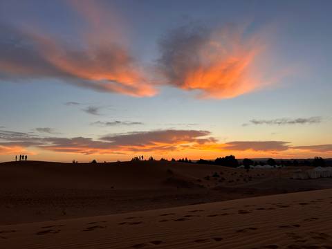 Fiery sunset clouds over golden Saharan dunes with silhouettes of travelers on the ridge.
