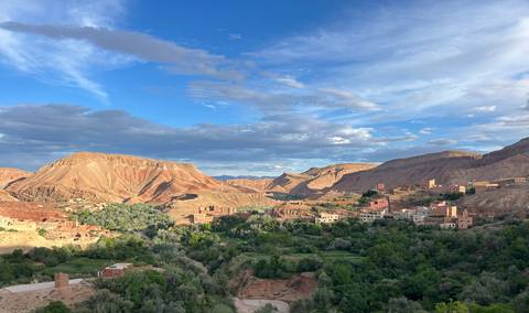 Wide valley of red cliffs, green oasis and scattered Berber villages under dramatic blue sky.