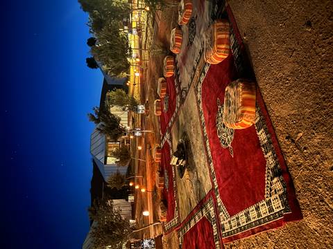 Atmospheric desert camp at night with carpets, fire pit and lanterns glowing under deep blue sky.