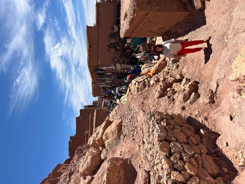 Market alley climbing the red-stone slope of Ait Benhaddou with craft stalls and blue sky.