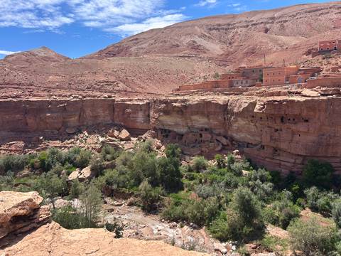 Red cliff walls with ancient cave dwellings above a lush green valley oasis.