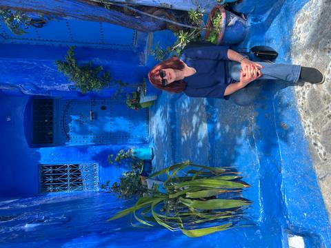Woman kneels by a vivid blue doorway decorated with potted plants in Chefchaouen.