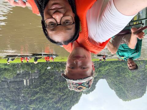 Selfie of two young travelers wearing life vests in a small rowing boat on a brown river surrounded by lush karst mountains and rice paddies, with other tourist boats and a local rower visible in the background.