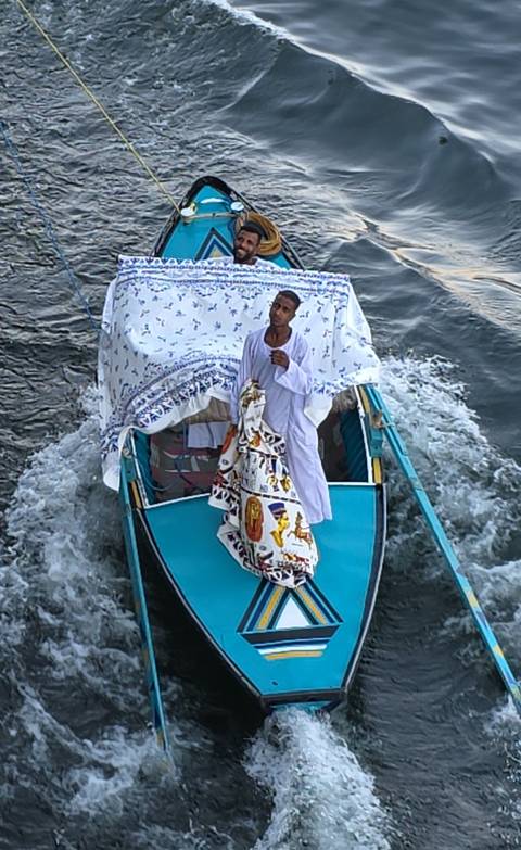Man in traditional white robe standing on the bow of a small boat decorated with patterned cloth on the Nile