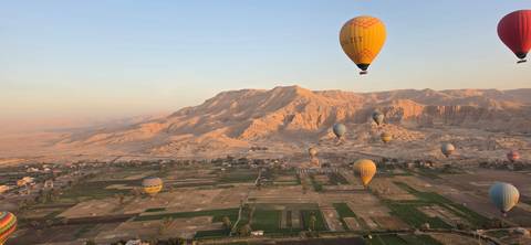 Hot-air balloons drift over patchwork fields and rugged cliffs of Luxor’s West Bank at sunrise.
