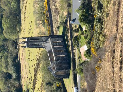Ruins of a stone church with a tall tower sitting in a green rural valley framed by hedges and fields.
