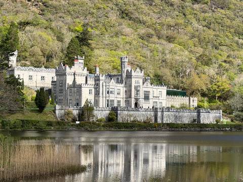 Kylemore Abbey’s grey-stone castle façade reflected in a calm lake at the foot of forested hills.