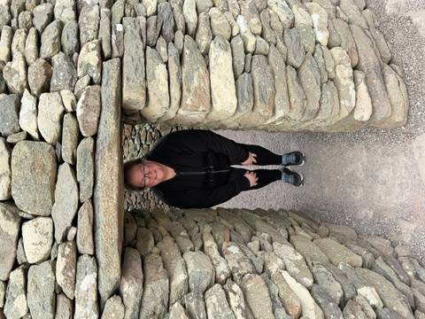 A traveller crouches and smiles inside a narrow doorway of an ancient dry-stone beehive hut.