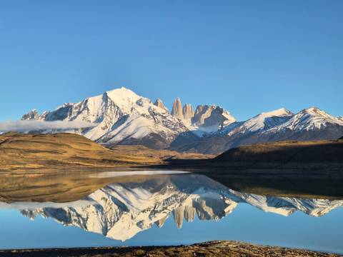 Snowy Torres del Paine massif perfectly reflected in a still lake at dawn, with golden foothills in the foreground.