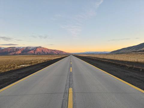 A long straight highway disappears into a pastel sunrise horizon between rugged Patagonian ranges.