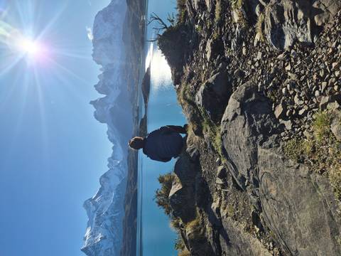 A hiker sits on a rocky outcrop admiring bright blue Patagonian lake waters with towering snowy peaks beyond.