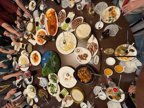 Round restaurant table crowded with plates of Chinese dishes, drinks and chopsticks during a lively shared meal.