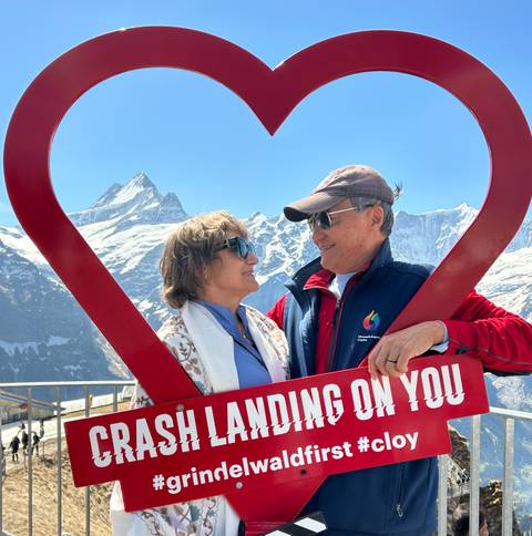 A smiling couple framed by a large red heart enjoy sweeping snowy alpine peaks under a clear sky.