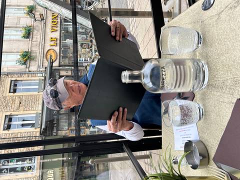 Seated at an outdoor café, a man studies a large menu with city buildings in the background.