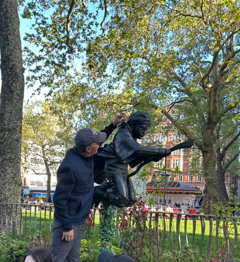 A tourist jokingly interacts with a bronze statue of a historical figure in a leafy square.
