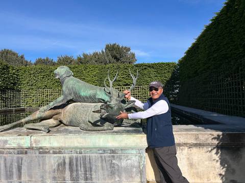 A visitor poses with a dramatic bronze hunting sculpture of dogs and deer in a manicured garden.