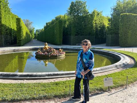 A traveller smiles beside a gilded fountain sculpture in a formal hedge-lined garden.