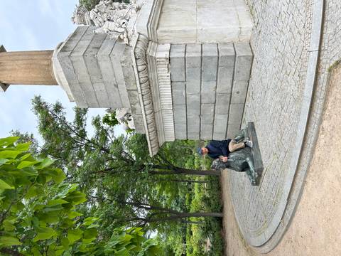 A man kneels playfully beside a bronze sphinx statue at the base of a stone monument.