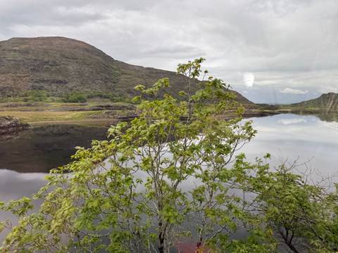 A reflective lake and rugged hills are seen through a bus window, faint reflections visible on the glass.