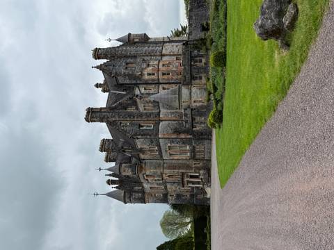 Large stone Victorian-style castle with turrets and chimneys set beside a manicured green lawn under an overcast sky.