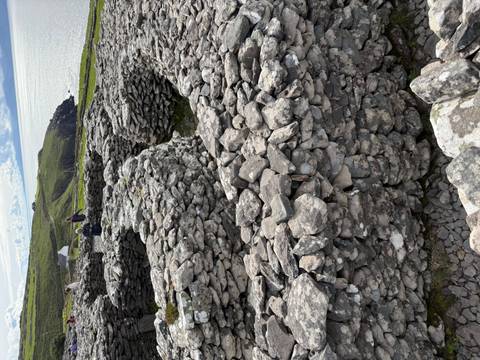 Ancient circular stone huts and walls overlooking green coastal hills with visitors exploring the archaeological site.