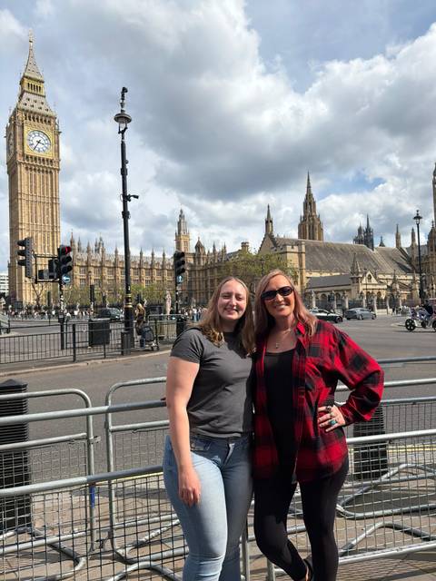Two travellers smile in front of London’s Houses of Parliament and Big Ben on a partly cloudy day.