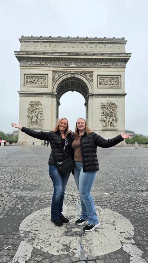 Two travellers with arms wide open in front of the Arc de Triomphe on an overcast Paris morning.