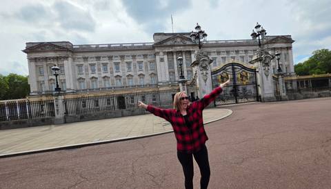 Happy traveller stretching arms before Buckingham Palace’s ornate gates and façade under dramatic clouds.