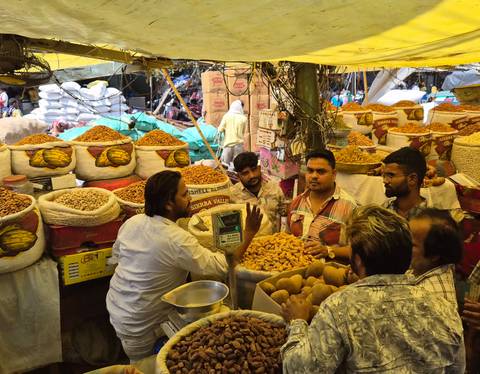 Bustling spice market stall in Delhi with vendors and sacks overflowing with nuts and dried fruit.